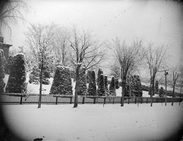 View from road towards a picket fence around a snow-covered yard with sculpted shrubs. The side of the house is on the far left. Identified as the Spaulding residence.