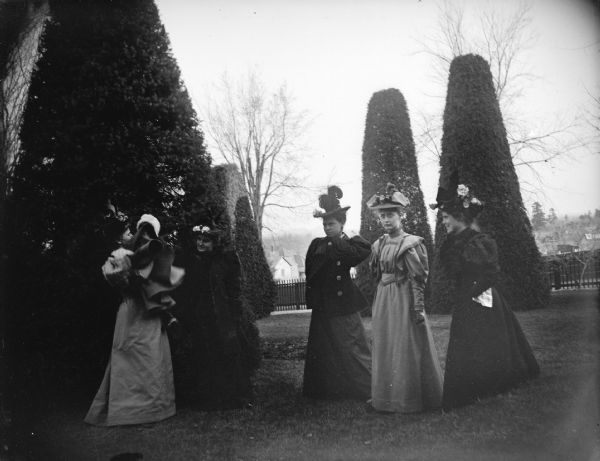 Outdoor group portrait of five European American women posing standing on a well-manicured yard among sculpted shrubs. The woman on the far left is holding a small girl in her arms. Identified as the Spaulding residence.
