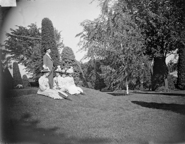 Outdoor group portrait of a man posing standing, and four women posing sitting on a slope on the lawn of a well-manicured yard with sculpted shrubs. Identified as the Spaulding family in the yard of their residence.