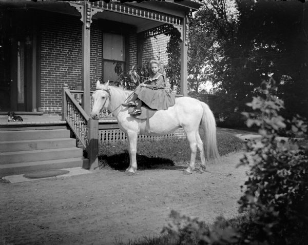 Outdoor portrait of a European American girl posing sitting side-saddle on a pony near the front porch of a brick building. A small dog is sitting on the porch at the top of the steps. Location identified as probably the porch of the Spaulding house.