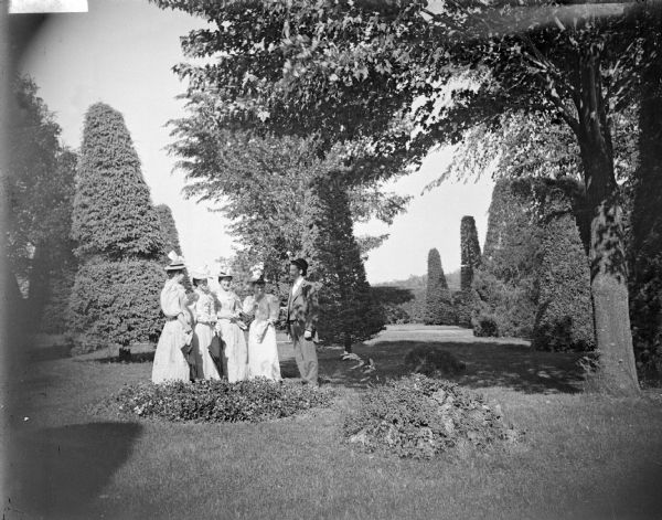 Outdoor group portrait of a man and four women posing standing on the lawn of a well-manicured yard with sculpted shrubs. Identified as the Spaulding family in the yard of their residence.