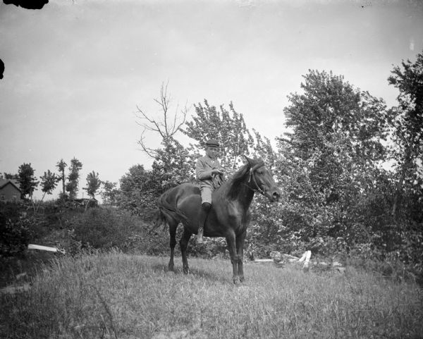Outdoor portrait of a boy posing sitting bareback on a horse in a field in front of some bushes. There is a building in the background.