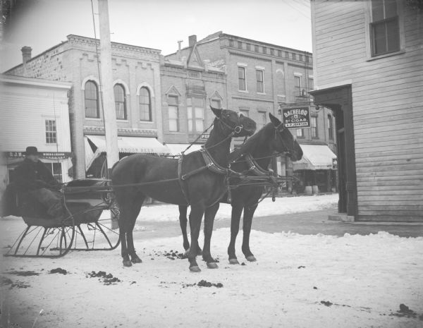 Outdoor view of a man posing sitting in a sleigh pulled by a team of two horses on a street in town. Location identified as the intersection of First and Main Streets.