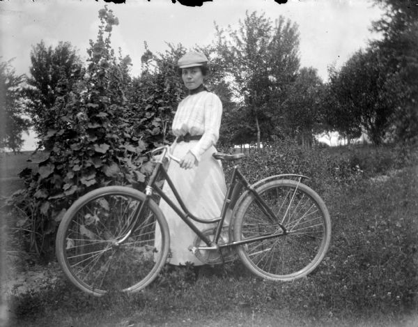 Outdoor portrait of a European American woman, wearing a long white dress, and a cap, posing standing near plants in a yard. She is holding a bicycle.