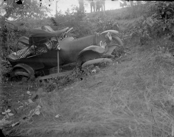 View looking uphill towards a wrecked automobile resting in a ditch off the road. Probably due to an accident. People are standing near the top of the hill looking down at the automobile.