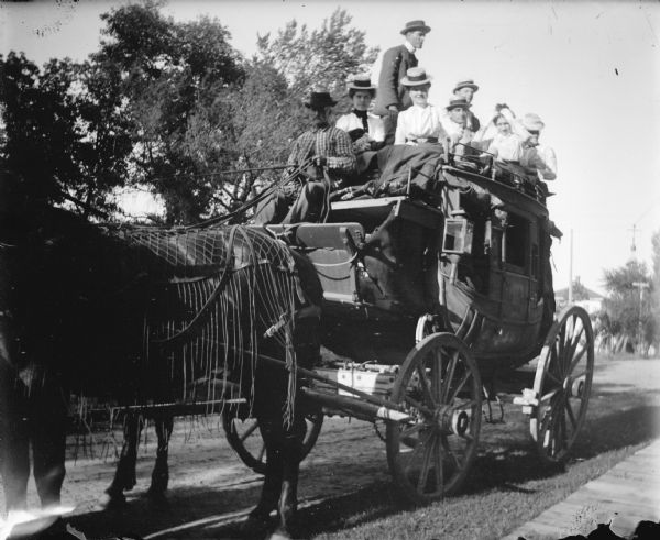 Outdoor group portrait of a stagecoach pulled by a team of two horses. A man is driving, and seven men and women are sitting and standing on top of the roof. Identified as the Price Stagecoach.