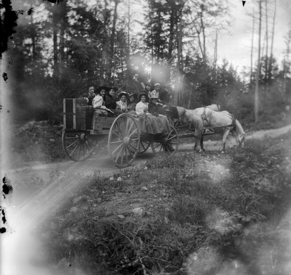 Outdoor group portrait from field towards a large group of men and women posing sitting in a wagon pulled by a team of two horses on an unpaved road.
