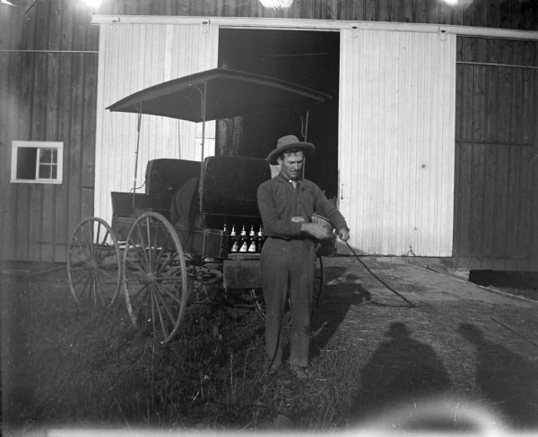 Outdoor view of a man posing standing in front of a surrey and a large barn with an open door. There are bottles stacked in the back of the surrey. The shadows of two men are in the foreground.