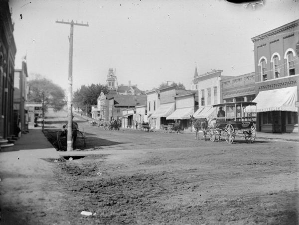 View from side of street towards a surrey pulled by a team of two horses moving down the street near storefronts. There are other horse-drawn vehicles further down the street.