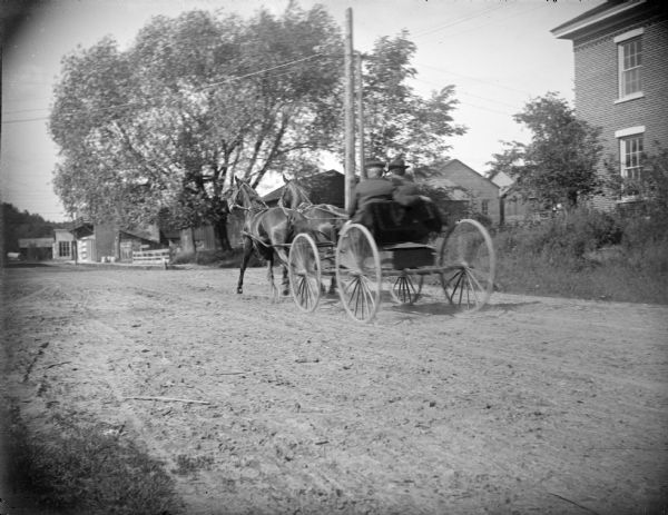 Exterior view from left rear of two men sitting in a wagon pulled by a team of two horses down an unpaved road. Location identified as probably looking north on Second and Fillmore Streets.