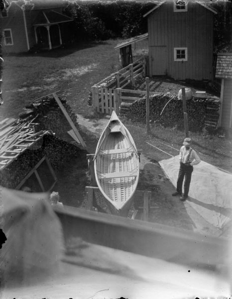 Elevated view of a man posing standing next to a canoe on sawhorses in a yard, showing the interior of the canoe, and two paddles lying inside. A young girl is standing to the left of the canoe. Fuelwood stacks are on the left and behind the man along a fence. There is a building in the background, and what may be a house across an open yard on the left.