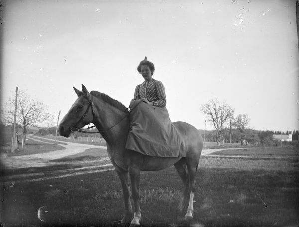 Exterior portrait of a woman posing sitting side-saddle on a horse in a field. There are roads in the background, and a building in the distance on the right.