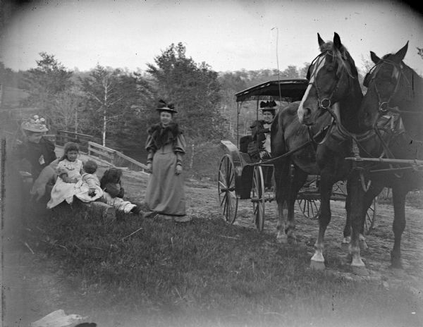 View towards three women and three children stopped on a roadside. One of the women is sitting next to the children in the grass on the side of the road. Another woman is standing next to them, and the other woman is posing sitting in a surrey pulled by a team of two horses. In the background are the wooden railings of a bridge.