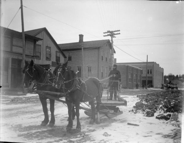 Three-quarter view from front left of a man posing standing on a heavy sled behind a team of two horses on a snow-covered street. Location identified as South First Street. There is a row of commercial buildings across the unpaved street, and a horse-drawn wagon in the background.