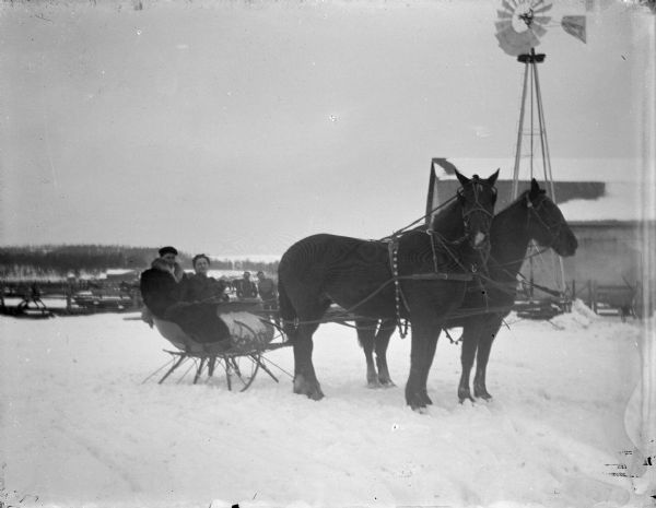 Outdoor view of a man and woman posing sitting in a sleigh pulled by a team of two horses on snow-covered ground. Two men are standing in the background behind the sleigh.