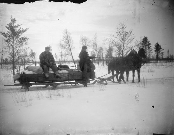 Outdoor portrait of two men posing sitting on a sled pulled by a team of two horses over a snow-covered field.