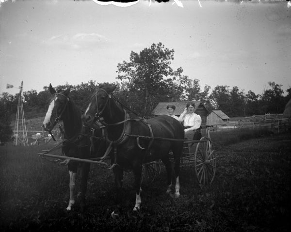 Outdoor portrait of two women posing sitting in a two-wheeled buggy behind a team of two horses. There is a windmill and farm buildings in the background.