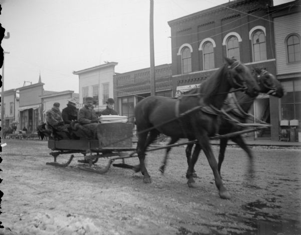 Exterior portrait of three men and a woman posing sitting in a sleigh being pulled by a team of two horses on a snow-covered road in town. Location identified as Main Street in Black River Falls.