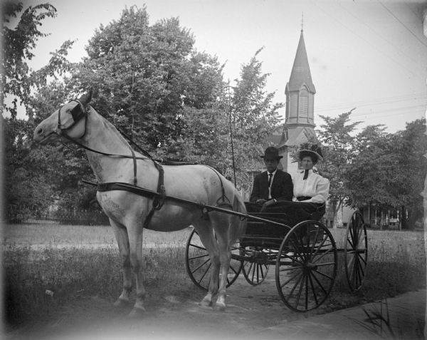 Exterior portrait of a man and woman posing sitting in a wagon pulled by a single horse. Location identified as in front of the Methodist Church, its steeple visible in the background.