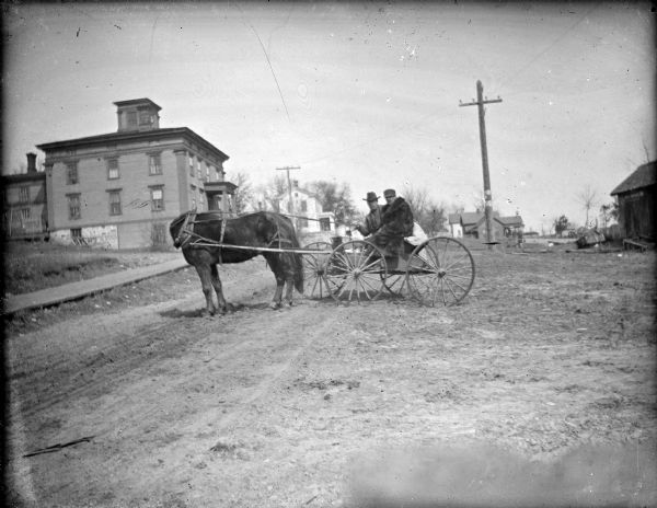 Outdoor view of two men posing sitting in a wagon pulled by a single horse on an unpaved road. The horse's head is turned sharply to the right. The building on the left is probably the Spaulding Boarding House.