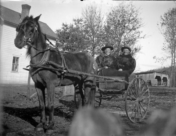 Outdoor view of two men posing sitting in a two-wheeled wagon pulled by a single horse. There are wooden buildings in the background.