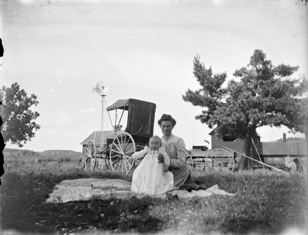 Outdoor portrait of a woman posing sitting and holding a child on a blanket in a field in front of two wagons, wooden buildings and a windmill.