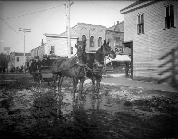 Outdoor view of a team of horses pulling two men in a sleigh, with an intersection in the background. The street is wet and muddy. Location identified as First and Main streets, looking north.