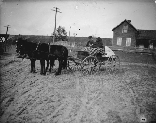Outdoor view of a man, woman and child, sitting in a wagon with a blanket over their laps, pulled by a team of two horses. In the background on the right is a wooden house behind a fence.