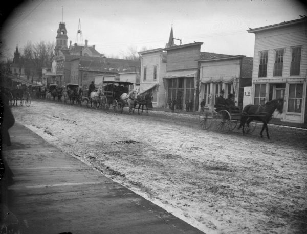 View from board sidewalk of a line of wagons, including a hearse, moving down an unpaved street in town. Location identified as Main Street, and a funeral procession.