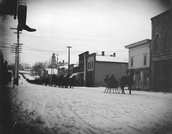 Outdoor view from side of snowy street of a long line of sleighs and wagons moving through the town center. Location identified as Main Street, and a funeral procession.