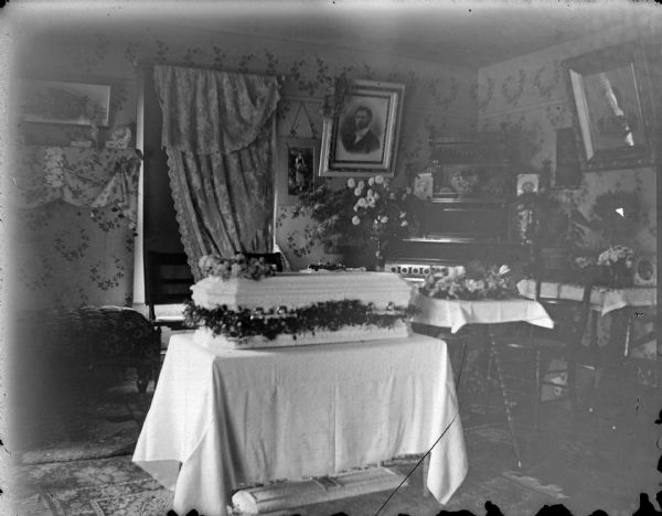 Indoor view of a small, light-colored casket resting on a table covered with a drape. The coffin is surrounded by flowers and flower arrangements on tables in a room with an organ in the corner, and several portraits of people hanging on the walls.
