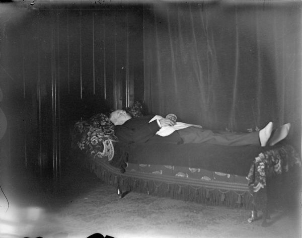 Interior postmortem portrait of a dead man lying on a bed in a room.