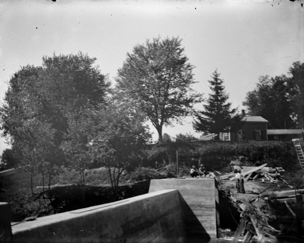 Outdoor view across dam on a river towards people on the far shoreline, with a building on the hill behind them. Location identified as the Dells dam near Hatfield.