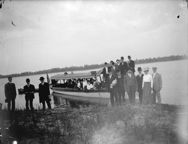 View from shoreline towards a large group of men and women posing on the shore, and on boat moored to the shore of a lake. Location identified as a 1908 boat ride on the lake.