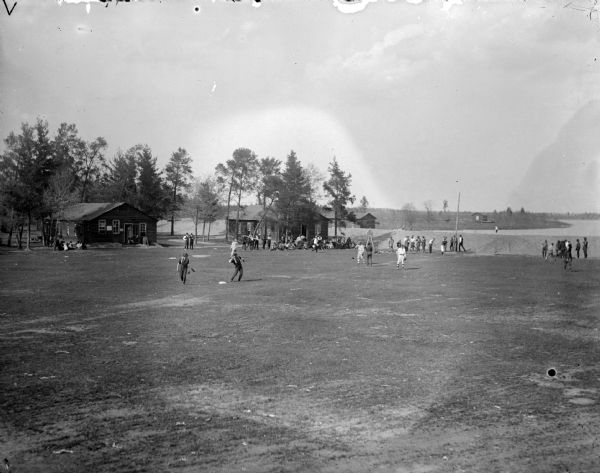 Outdoor view across field towards men playing baseball. There is a small crowd sitting and watching near two wooden buildings on the far side of the field. Location identified as Hatfield.