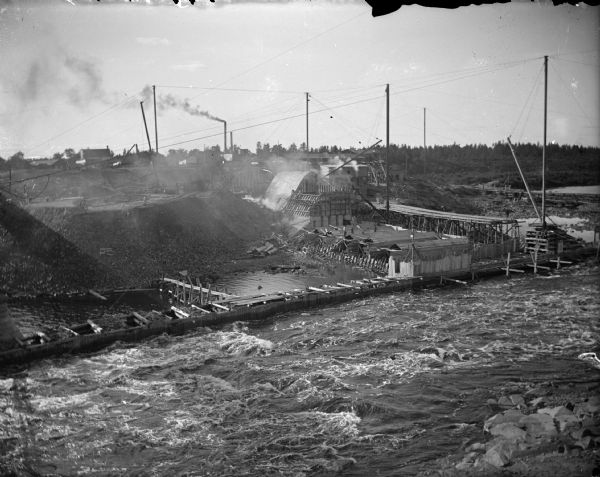 Elevated view across rushing water towards a construction site of a dam on a river. Identified as the construction of the Hatfield Dam, looking northwest.
