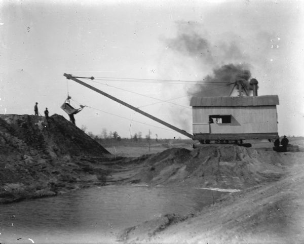 View of a steam dredge at the construction site of a dam on a river. Identified as the construction of the Hatfield dam in 1907.
