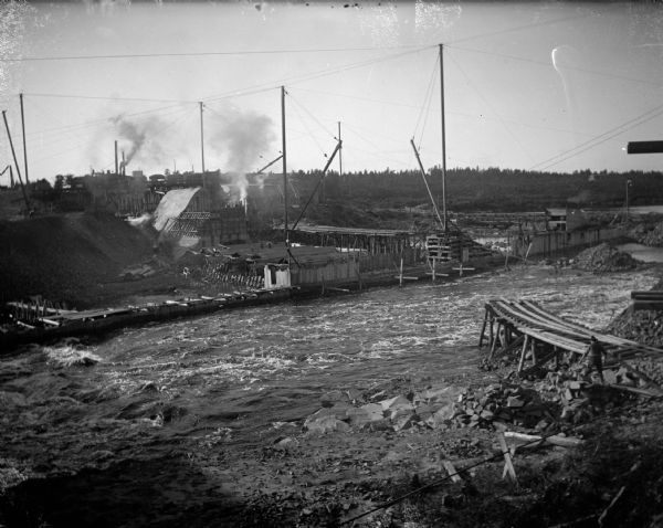 Exterior portrait of the construction site of a dam across a river. Identified as the construction of the Hatfield dam across the Black River looking west in 1907.