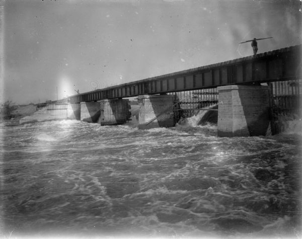 View across water looking up at a railroad bridge over a river. A man is walking across the bridge while carrying what may be lumber. Identified as the railroad bridge behind the gates of the Hatfield dam.