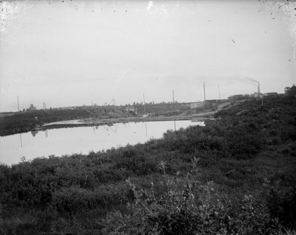 View over shrubs and plants towards the construction site of a dam across a river. Identified as the construction of the Hatfield dam across the Black River in 1907.