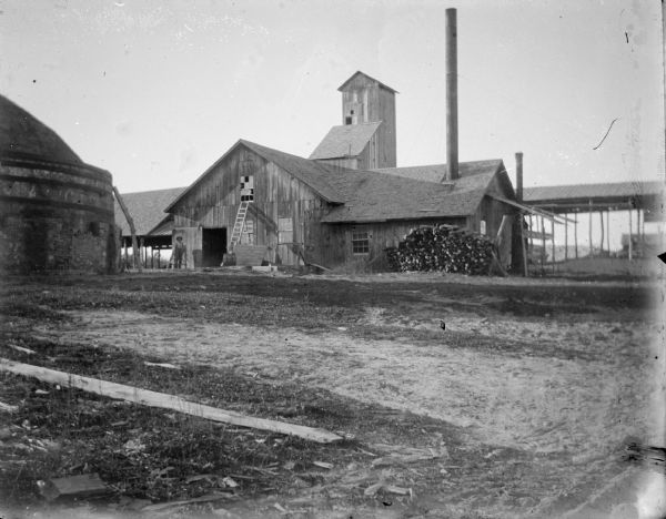 Outdoor view across yard towards a man posing standing by a group of buildings, including a tall wooden building in the background. There is a large kiln on the left. Identified as the yard at the Halcyon Brick Works.