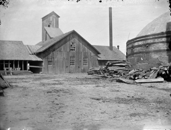 View across yard towards buildings, including a tall wooden building. On the right is a large kiln, with fuel wood stacked in front. Identified as the yard at the Halcyon Brick Works.