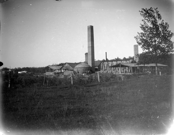 View across field towards tall chimneys, wooden buildings, and a large kiln. Identified as the Halcyon Brick Works, approximately halfway between Black River Falls and Hatfield.
