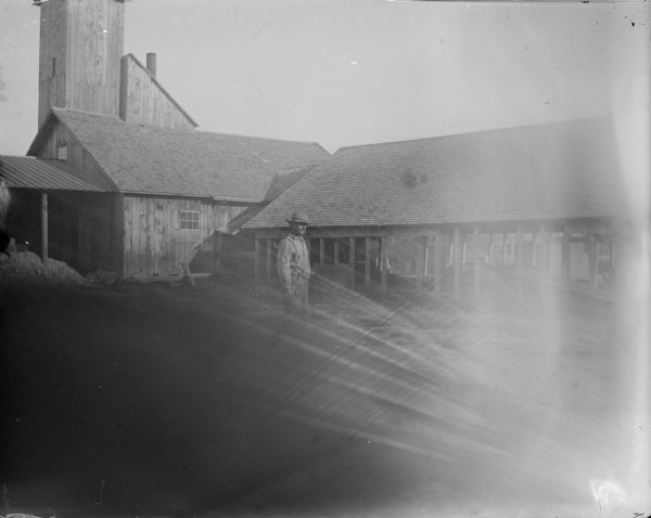 View across yard towards a man posing standing near buildings, including a tall wooden building. Identified as the yard at the Halcyon Brick Works.