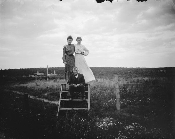 Outdoor group portrait of a man posing sitting, and two women posing standing on a fence ladder in a field. The Halcyon Brick Works is identified in the background.