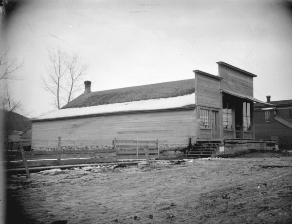 View across unpaved road towards storefronts on a main street. Identified as Abbott's Grocery Store on the northwest corner of the Main Street intersection in Hixton.