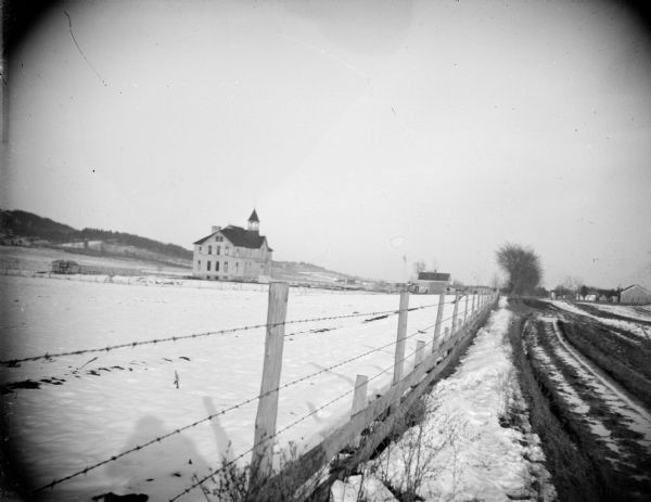 View along fence line snow-covered field towards a three story building in the distance on the left. Identified as the Hixton school towards Sechlerville.