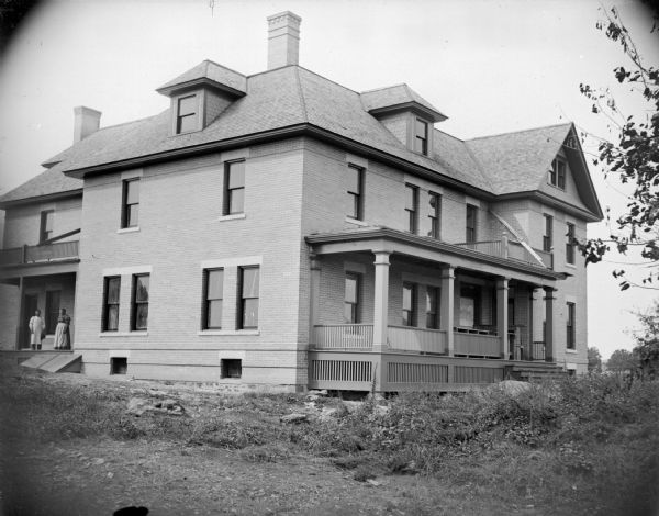 View across yard towards a large two-story brick house with front porch. There is a man and woman posing standing on another porch on the left. Identified as a house just north of Hixton towards Northfield.