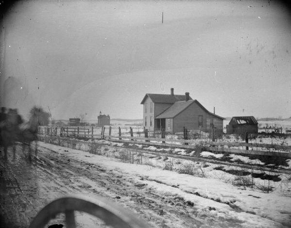 View across road towards a house and outbuildings, and a school building in the distance, in snow covered fields. On the left is a wagon coming up the muddy road pulled by two horses. Identified as outside Hixton, and the school building on the edge of Hixton.