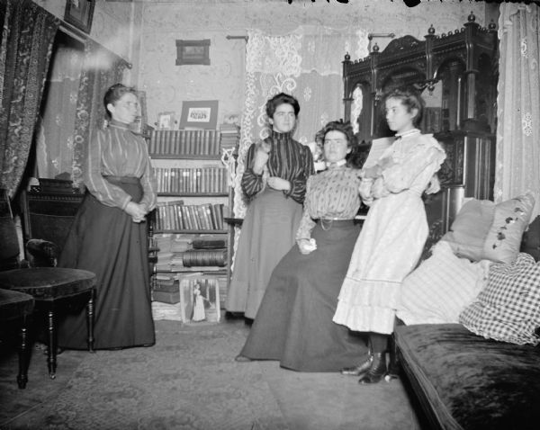 Interior portrait of four women posing sitting and standing in a room next to an organ and a book shelf.